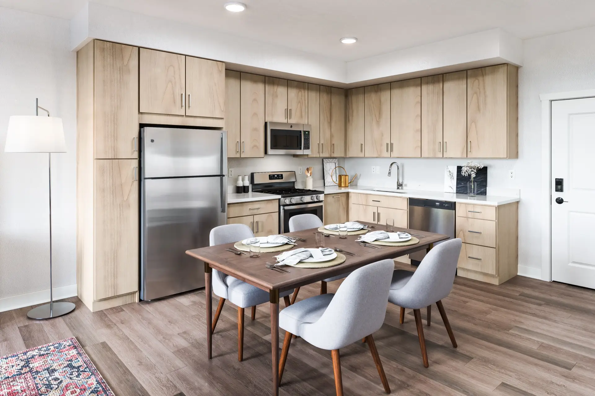 Modern kitchen with light wood-style cabinets, stainless appliances, dining table for four with gray chairs, and bright neutral decor.