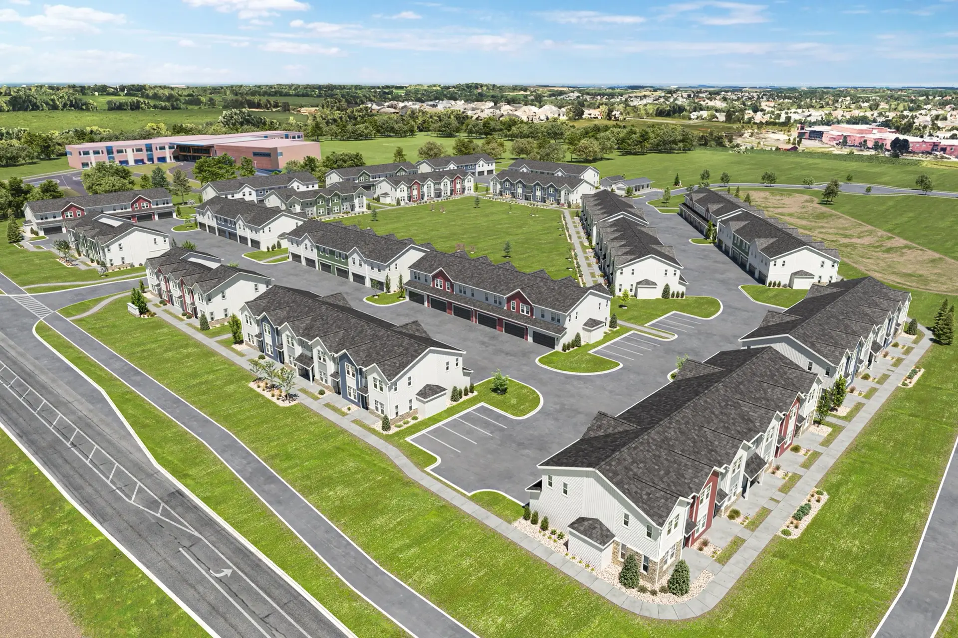 Aerial view of a modern townhouse complex with gray buildings, green lawns, parking, roads, open fields, and distant buildings under blue sky.