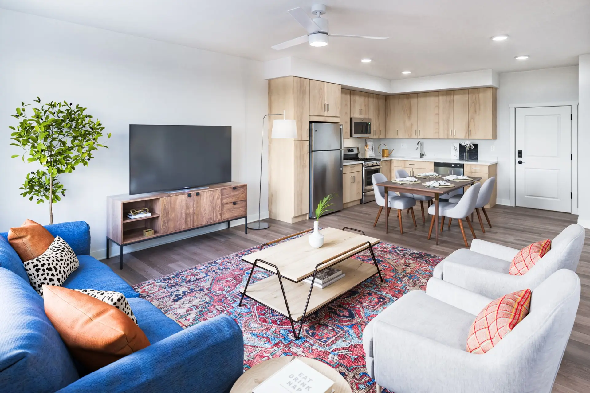 Modern open-plan living room and kitchen with wood-look cabinets, stainless appliances, blue sofa, gray chairs, colorful rug, TV stand.