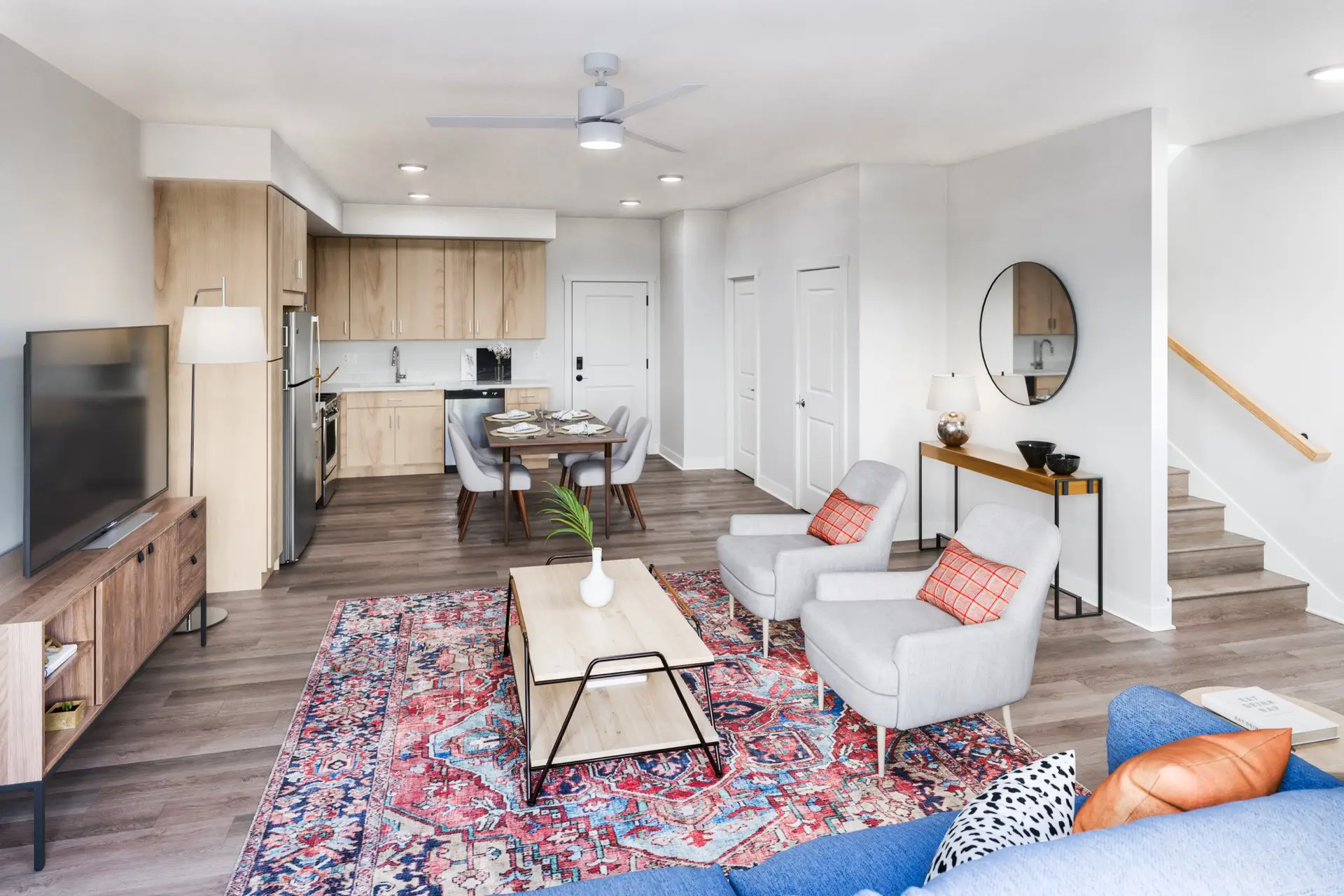 Modern open-concept living room and kitchen with wood-style floors, light wood-style cabinets, colorful rug, gray sofas, and dining table.