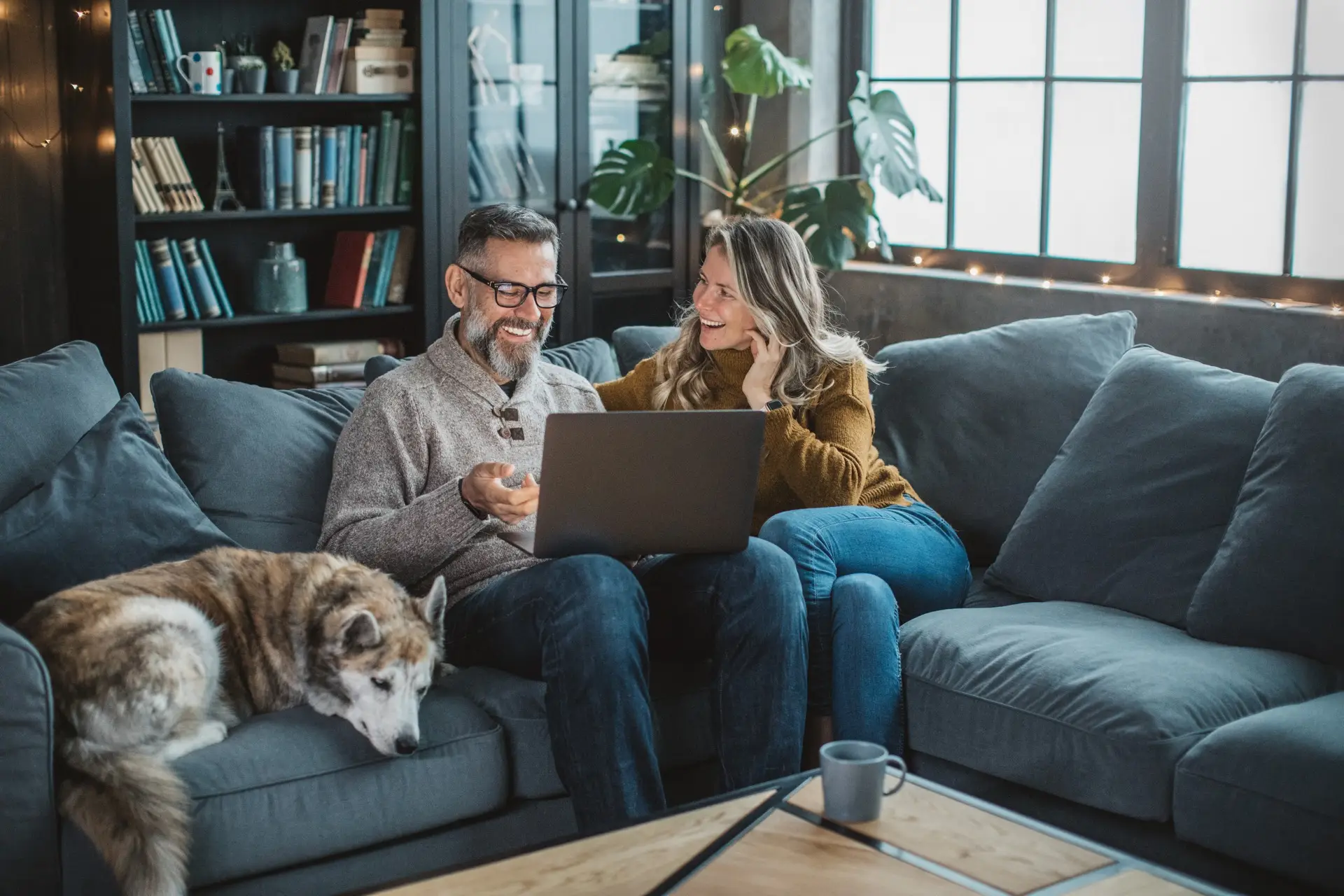 A smiling man and woman sit on a couch, looking at a laptop. A dog rests nearby. Wood-style shelves and string lights warm the cozy room.