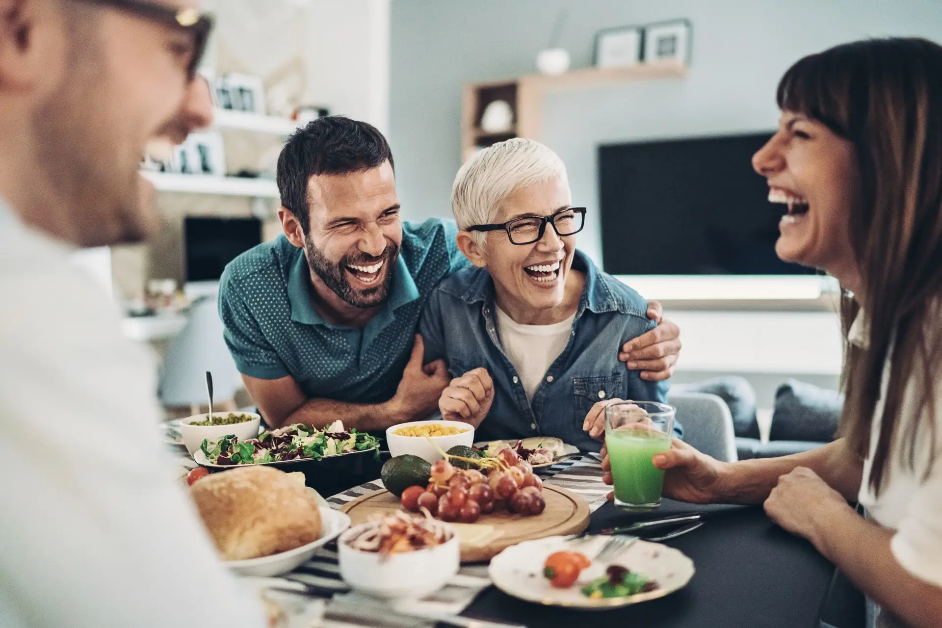 Four adults sit around a wood-look table, laughing and enjoying a meal with various dishes, salads, bread, and drinks. Warm atmosphere.