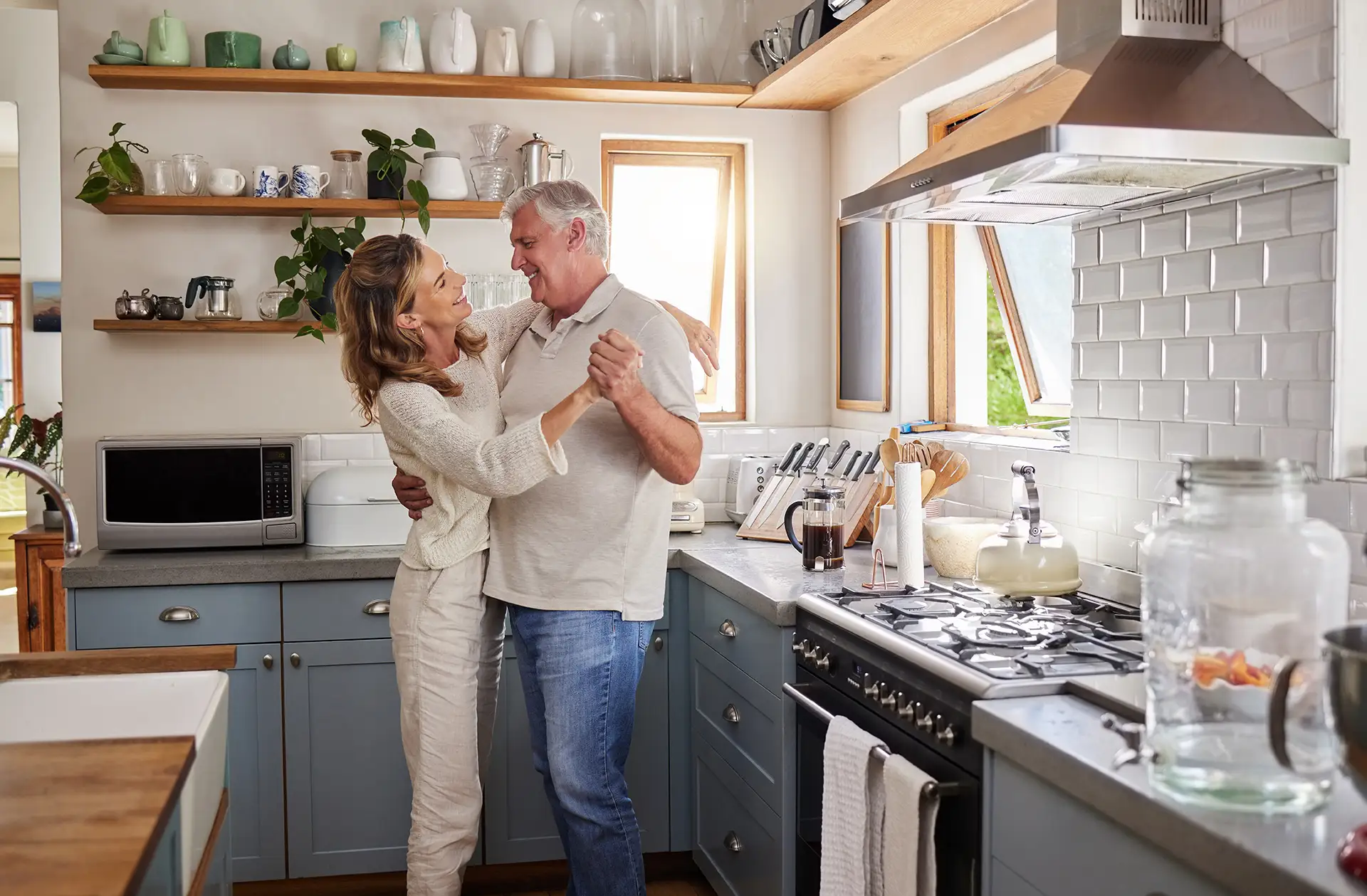 An older couple smiles and dances in a cozy, sunlit kitchen with blue cabinets, plants, and wood-style shelves, creating a joyful vibe.