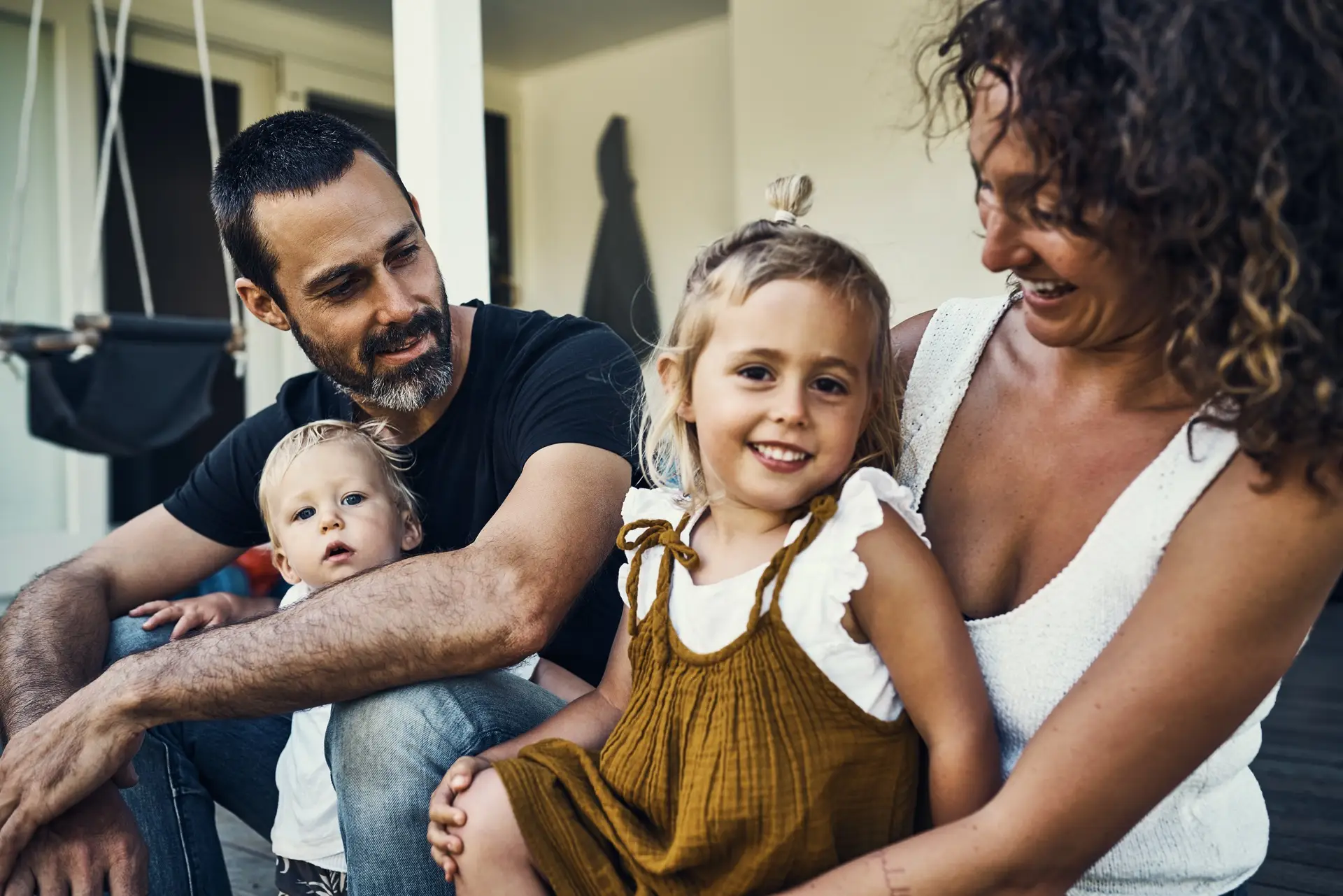 A family of four sits outside on a wood-like porch, smiling together. A man holds a young child while a woman hugs an older girl.