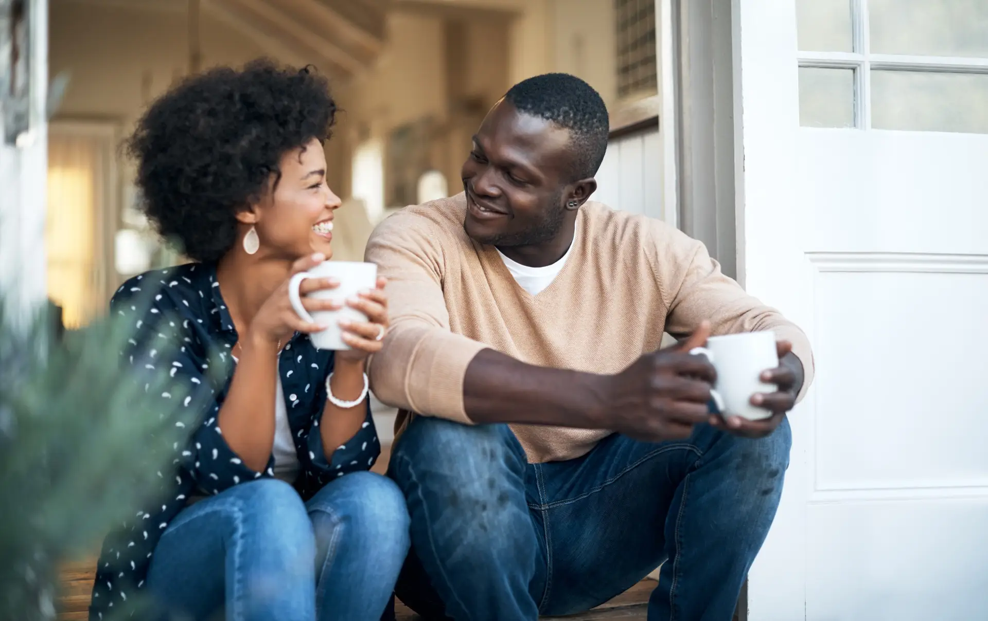 A smiling couple sits together on a wood-look porch, holding mugs and sharing a relaxed, happy moment while enjoying each other's company.