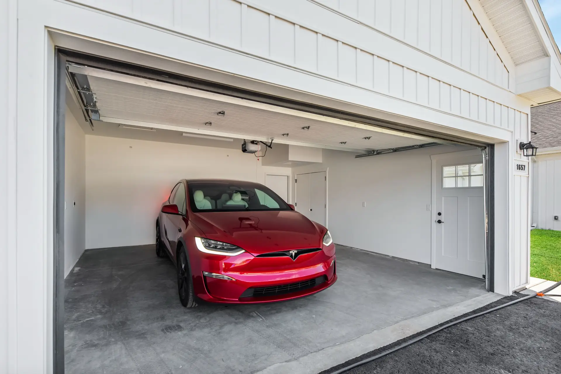 A red Tesla is parked in a clean, modern two-car garage with white walls and an open garage door. Grass is visible outside.